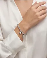 Pulsera elástica de acero compuesta por pequeñas esferas metálicas. Presenta dos dijes colgantes: uno en forma de corazón y otro rectangular con la inscripción 'Good Luck'.