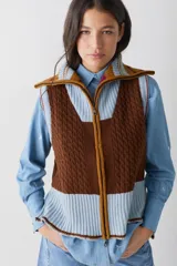 Chaleco de lana con cierre frontal completo y cuello alto. Presenta un diseño de bloques de color en tonos marrón chocolate y celeste, combinando secciones de tejido trenzado con secciones de tejido acanalado. Posee un corte asimétrico, siendo más largo en la parte posterior, y aberturas laterales en el ruedo.