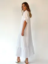 Vestido largo camisero de lino blanco, con cuello clásico, mangas cortas con presilla y botones, cierre frontal con botones y falda con volantes.