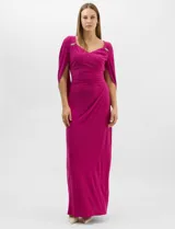 Vestido largo de seda fría color fucsia, con escote cruzado drapeado y apliques de pedrería en los hombros. Presenta mangas largas abiertas estilo capa y un drapeado lateral en la cintura que estiliza la silueta.