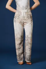 Pantalón de corte recto y tiro alto, de color beige con un estampado paisajístico de árboles y naturaleza en tonos marrones y grises. Cuenta con trabillas en la cintura y cierre frontal oculto.