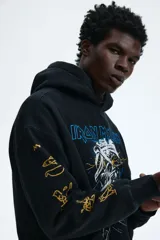 Canguro negro de corte oversized con capucha, bolsillo frontal tipo canguro y estampado frontal de la banda Iron Maiden con diseño de momia. Las mangas presentan detalles estampados en color dorado con motivos egipcios.