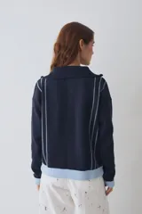 Buzo de tejido de punto color marrón con cuello alto y cierre frontal parcial. Presenta rayas verticales finas en color claro, puños y ruedo en tejido acanalado color crema, y detalles en color verde lima en el tirador del cierre, el cordón de ajuste del ruedo y el parche bordado en el pecho.