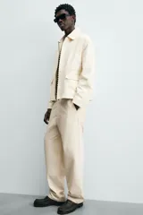 Pantalón chino de algodón color beige, de corte relajado (relaxed fit) y tiro medio. Presenta bolsillos delanteros y bolsillos traseros de vivo con cierre de cremallera y botón.