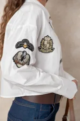 Camisa blanca de algodón, de corte corto y silueta holgada. Presenta cuello clásico, cierre frontal con botones y mangas largas con puños anchos. Está decorada con diversos parches y pines metálicos de estilo militar y náutico distribuidos en el frente y la manga derecha.