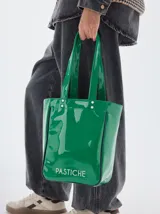 Bolso tipo tote de charol en color verde brillante, con doble asa de mano y detalle de ribetes en contraste. Presenta el nombre de la marca estampado en la parte inferior frontal y remaches metálicos en la base de las asas.