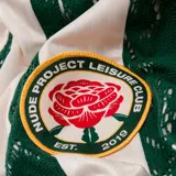 Camiseta de manga larga estilo jersey de fútbol, con diseño a rayas verticales alternando blanco y encaje verde. Presenta cuello polo con detalles en blanco y verde, y un escudo circular bordado en el centro del pecho con una rosa.