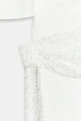 Vestido corto blanco con mangas cortas amplias y cuello recto. Presenta un fajín drapeado de lentejuelas plateadas a la cintura con un lazo largo lateral.