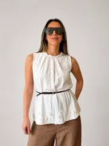 Blusa blanca sin mangas, con cuello trenzado fruncido, detalles de volados en el ruedo y un cinturón fino marrón incorporado.