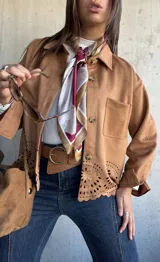 Chaqueta de gamuza color camel con cierre frontal de botones, cuello clásico y detalle de calados decorativos en el ruedo. Presenta bolsillos frontales y mangas largas con puños.