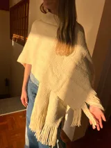Ruana color crema tejida en punto grueso con cuello alto y flecos en el borde inferior.