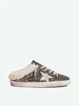 Zuecos Golden Goose modelo Super-Star Sabot, con diseño de zapatilla sin talón. Presentan exterior cubierto de glitter negro y dorado, estrella característica en el lateral, cordones y forro interior de shearling sintético.