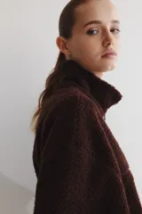 Campera oversized de sherpa suave color marrón oscuro, con cuello elevado, cierre frontal y bolsillos discretos en las costuras laterales. Presenta hombros caídos y mangas largas amplias.