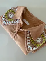 Camisa tostada de manga larga con cuello clásico decorado con apliques de crochet en tonos marrones, blancos y verdes, cierre frontal de botones y diseño holgado.