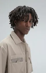 Camisa de manga corta color beige, estilo técnico, confeccionada en microfibra liviana. Cuenta con doble bolsillo frontal, uno con detalle de malla y otro con cierre, y broches en lugar de botones.