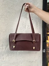 Bolso de hombro rectangular color marrón oscuro (ciruela/vino), con textura perforada y solapa frontal. Presenta dos hebillas decorativas doradas en la parte delantera y una correa delgada para llevar al hombro.