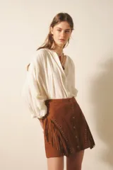 Falda corta de cuero gamuzado color marrón, con diseño de flecos laterales, cierre frontal con botones a presión y detalle de tachas metálicas y remaches decorativos.