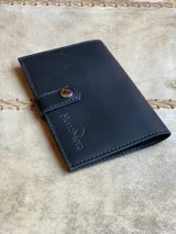 Billetera y porta documentos de cuero negro, con cierre de lengüeta con botón a presión y logo grabado de la marca Sierra Mora.