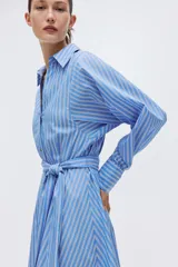 Vestido midi camisero de rayas azules y blancas, con cuello clásico, mangas largas, cierre frontal de botones y lazo ajustable en la cintura.