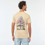 Remera beige de manga corta y cuello redondo, con pequeño estampado circular en el centro del pecho que incluye texto en tonos rosados y blancos.