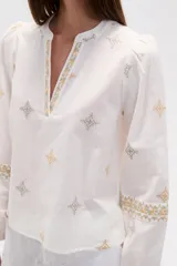 Blusa de algodón color tostado con cuello mao y escote en V. Presenta mangas largas abullonadas con puños elásticos y un diseño con bordados geométricos en contraste distribuidos por toda la prenda.