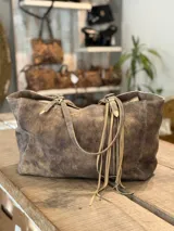Cartera tipo tote de cuero con efecto desgastado, color marrón arena. Presenta doble asa de hombro con detalles de hebillas metálicas y flecos largos decorativos en uno de los laterales.