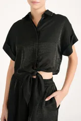 Camisa crop negra de tejido fluido y acabado brillante, con cuello camisero, mangas cortas con vuelta y nudo frontal en el dobladillo.