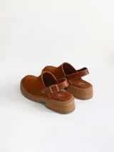 Zueco de cuero gamuzado color óxido, con tira trasera ajustable mediante hebilla metálica. Presenta una suela de plataforma con textura acanalada y detalle de textura reptil en la tira posterior.