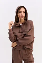 Hoodie de algodón color marrón con capucha, cierre frontal parcial y detalle de costuras horizontales en contraste negro. Presenta diseño holgado, mangas largas con puños elásticos y cordones ajustables en la capucha.