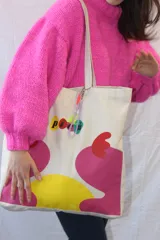 Tote bag de lienzo color crudo con estampado de diseño abstracto en colores rosa, amarillo y rojo. Incluye charm colgante con cuentas de colores.