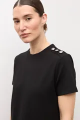 Vestido midi negro de manga corta y cuello redondo, con detalles de botones plateados en el hombro izquierdo, tajo lateral y un cinturón fino ajustable con hebilla metálica.
