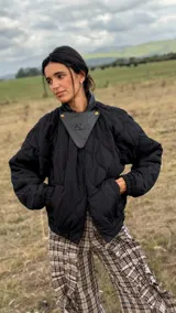 Campera puffer negra con diseño matelaseado en rombos, silueta amplia y corte bomber. Presenta cuello alto, cierre frontal y un detalle triangular de cuero en la parte superior con logo grabado. Incluye mangas largas con puños elásticos y bolsillos laterales funcionales.