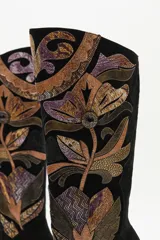 Bota de caña alta confeccionada en gamuza negra, con un diseño artesanal de parches en cueros texturizados y metalizados con motivos florales. Presenta un taco cónico y punta almendrada.