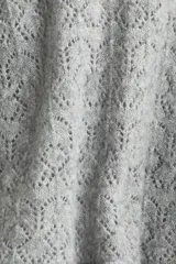 Buzo corto de punto pointelle color gris, con cuello redondo, mangas largas y terminaciones festoneadas en cuello y puños. Presenta un ribete acanalado con volados en el bajo.