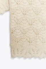 Jersey de punto abierto color crema, con cuello subido y mangas cortas abullonadas. Presenta un patrón de tejido calado con motivos en forma de hoja o escama y un borde festoneado en el ruedo.