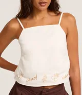 Blusa confeccionada en sarga de algodón, de color blanco, con escote cuadrado y tirantes finos. Presenta un detalle bordado en la parte inferior con motivos botánicos y la palabra ciao.