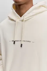 Buzo con capucha de corte holgado, color crema, con bolsillo frontal tipo canguro y pequeña estampa tipográfica en el pecho.