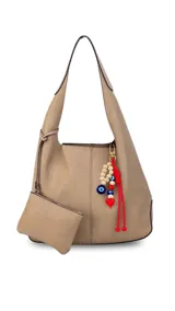 Cartera tipo hobo de cuero sintético en color beige, con doble asa de hombro integrada, costuras frontales visibles y un monedero colgante a juego con cierre. Incluye un colgante decorativo lateral con cuentas de madera, cordones rojos y dijes de mal de ojo y corazón.