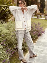 Cardigan de punto color beige con escote en V, cierre frontal con botones, bolsillos delanteros aplicados y detalle de flecos en el ruedo.