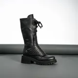 Borcego de cuero vacuno color negro, con suela de goma artesanal, caña alta con aplique capitoneado desmontable y horma de punta redonda.