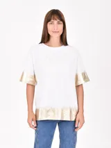 Remera blanca de corte holgado con detalles metalizados en color dorado en las mangas y el ruedo. Presenta un estampado minimalista con texto en el frente.