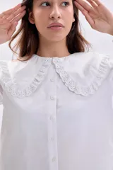 Camisa blanca de manga larga con cuello grande estilo babero con detalles de bordado inglés (broderie), cierre frontal con botones y puños elásticos con volados.