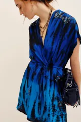 Vestido corto azul con estampado tie-dye en tonos negros y azules, escote en V cruzado, mangas cortas y cordón ajustable en la cintura. Presenta detalles de aplicaciones bordadas con lentejuelas en hombros y falda.