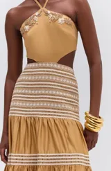 Vestido largo color mostaza, con diseño cut-out en la cintura, cuello halter con tiras para atar, y detalles bordados en el escote. La falda larga presenta paneles fruncidos con detalles tejidos en hilo crudo.