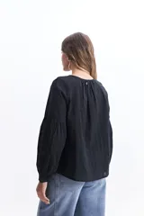 Blusa de manga larga color negro, confeccionada en 100% algodón con textura bordada. Presenta detalles de tablas en el frente y en los puños, con un corte holgado y cuello redondo con pequeña abertura.