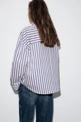 Camisa de manga larga con cuello subido y trabillas en los hombros. Presenta un diseño de rayas verticales en tonos azul, beige y marrón, con cierre frontal de botones.
