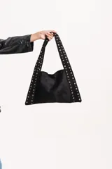 Cartera tipo tote bag de gamuza negra, con diseño triangular y correas integradas decoradas con tachas metálicas doradas en todo su contorno.