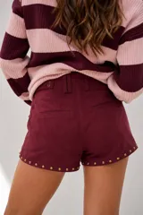 Short de tiro medio con efecto gamuza en color bordó, decorado con tachas metálicas doradas en los bordes y bolsillos. Cuenta con presillas para cinturón y cierre frontal.