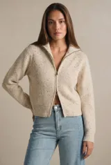 Cardigan de punto acanalado en color beige melange con motas oscuras. Presenta un diseño ajustado al cuerpo, cierre frontal completo con cremallera metálica y cuello alto.
