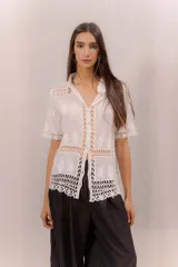Camisa blanca de manga corta con detalles de encaje calado y transparencias, cuello camisero y cierre frontal de botones.